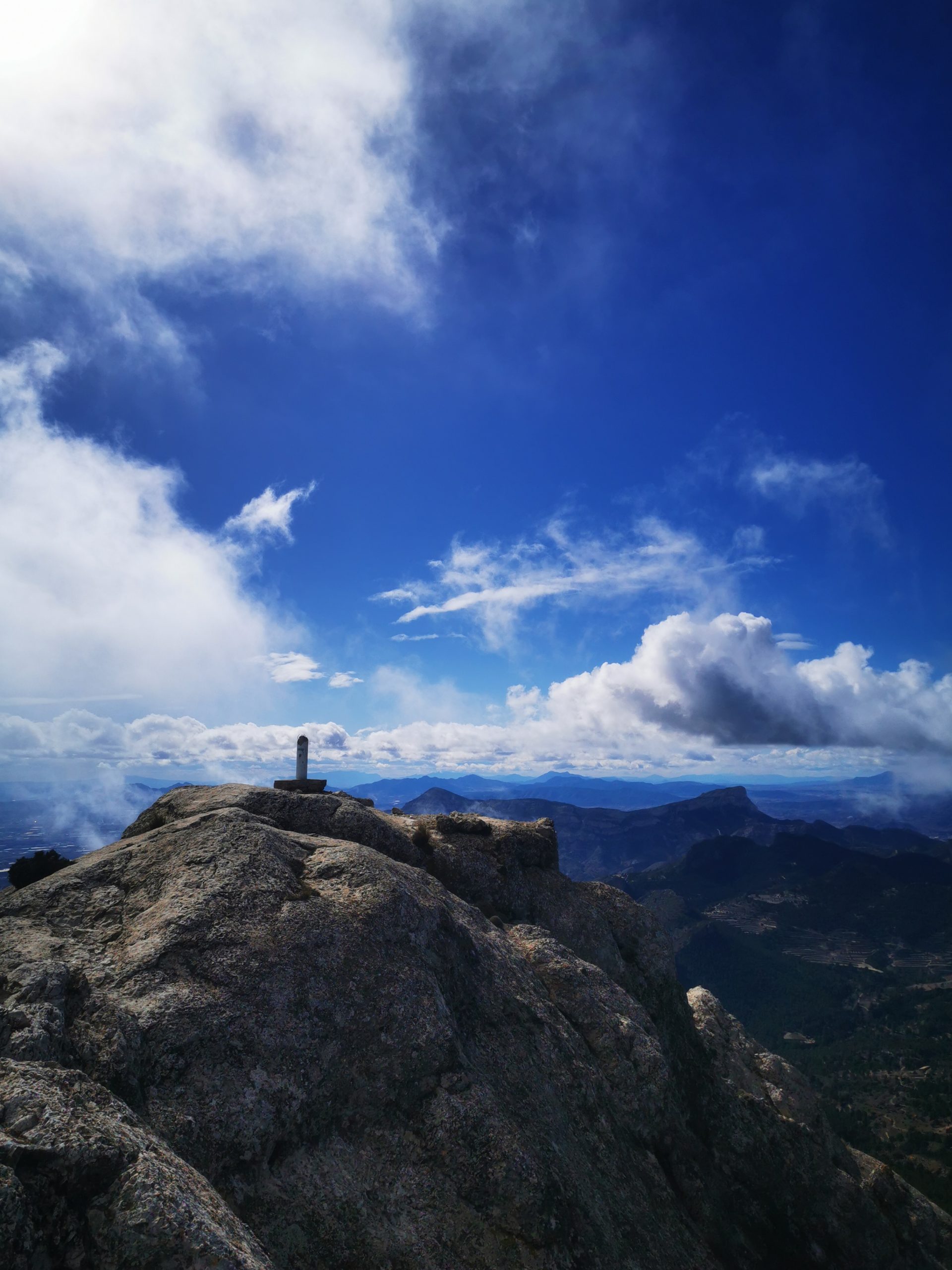 Ein felsiger Berggipfel unter einem weiten blauen Himmel mit entfernten, in Wolken gehüllten Bergketten.