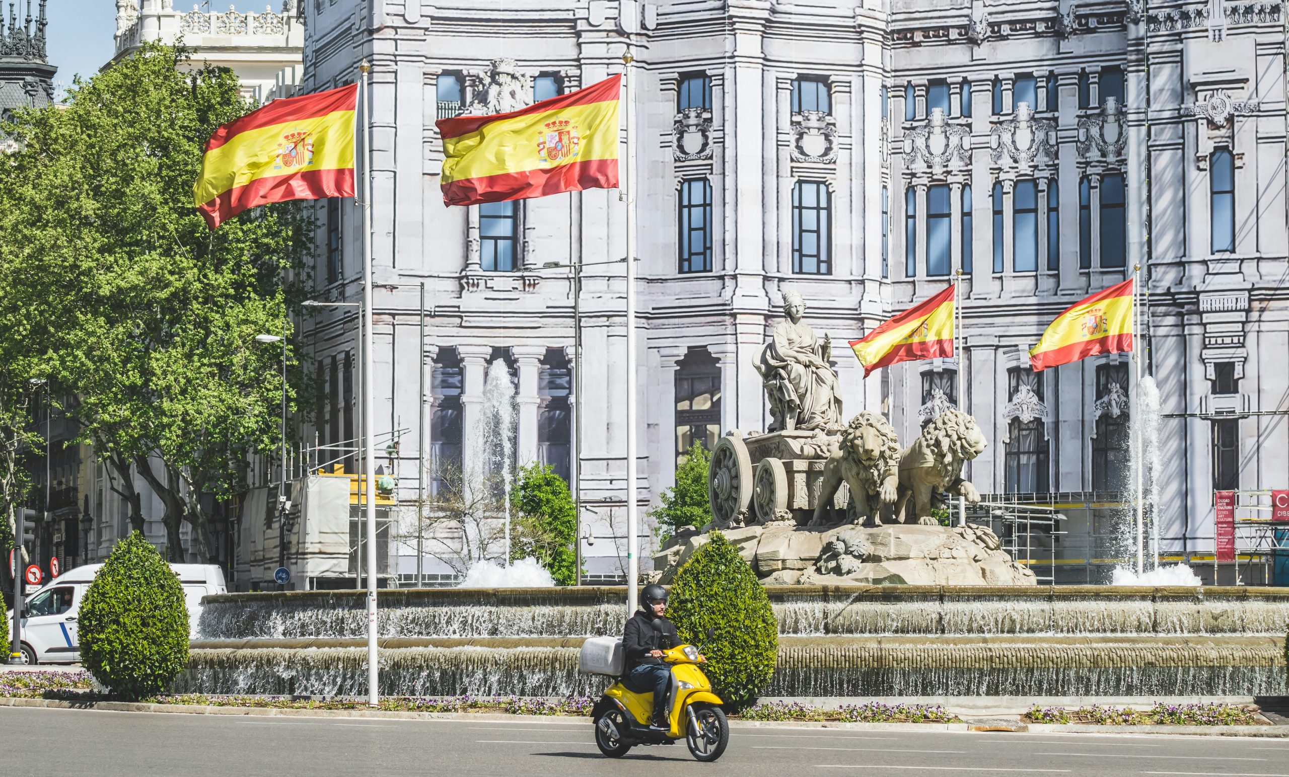 Ein gelber Motorroller fährt an einem Brunnen und einem Löwendenkmal vorbei, vor einem großen historischen Gebäude wehen spanische Flaggen.